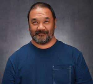 A swarthy man with close-cropped dark hair with salt and pepper beard wearing a blue t-shirt