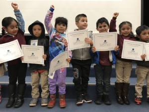 A group of young school children holding their Oakland Promise Certificates of Scholarship and cheering