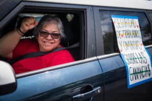 A grey haired smiling woman in a read shirt saluting from the window of a dark blue car. A solidarity sign is taped to the passenger window behind her.