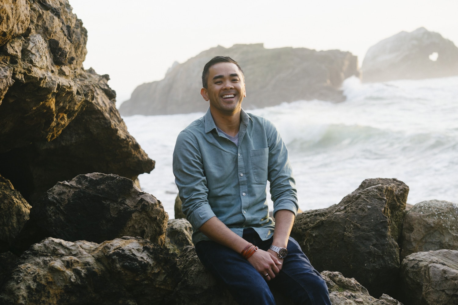 A photograph of a smiling man with short dark hair wearing a blue shirt and dark pants sitting on rocks at the ocean's edge