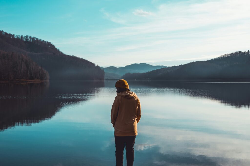 Person in yellow jacket standing near lake with mountains in the background