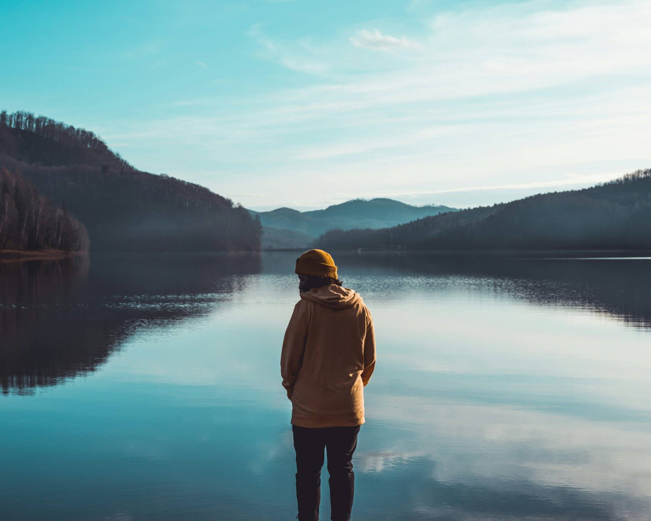Person in yellow jacket standing near lake with mountains in the background