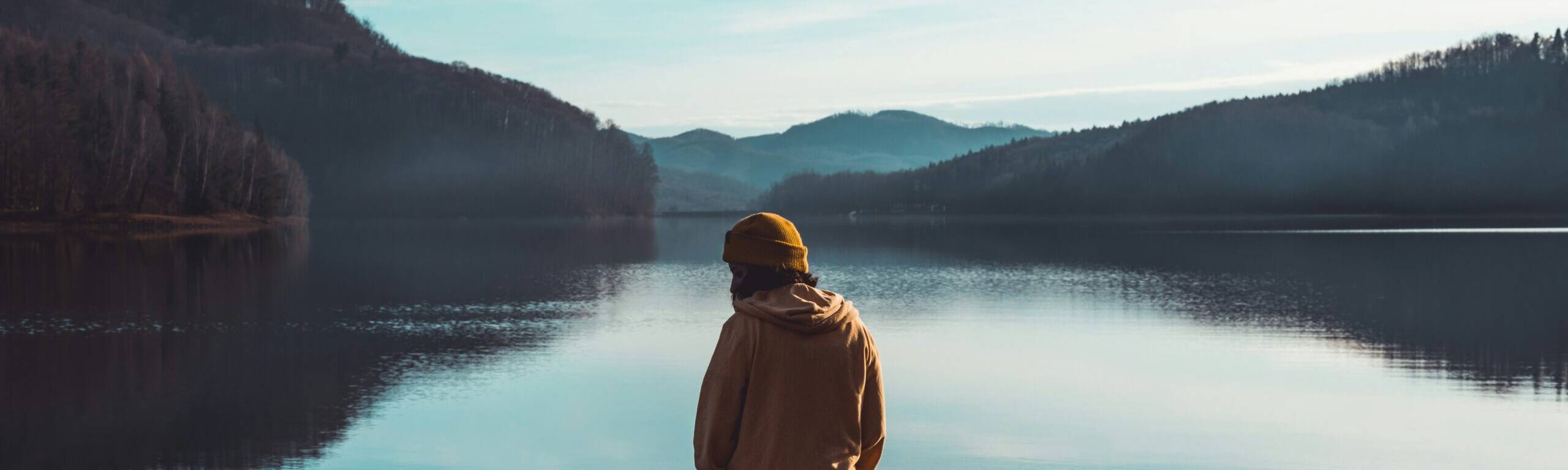 Person in yellow jacket standing near lake with mountains in the background
